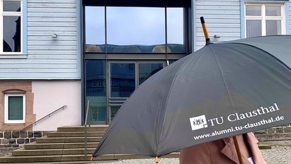 Person mit einem schwarzen Regenschirm mit der Aufschrift TU Clausthal vor dem Haupteingang des TU Clausthal Hauptgebäudes