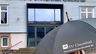 Person mit einem schwarzen Regenschirm mit der Aufschrift TU Clausthal vor dem Haupteingang des TU Clausthal Hauptgebäudes